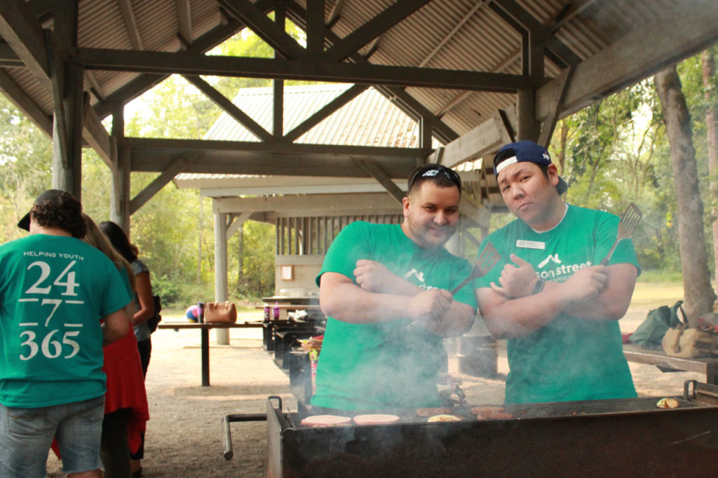 Two staff members smiling with arms folded and with spatulas over a BBQ. 